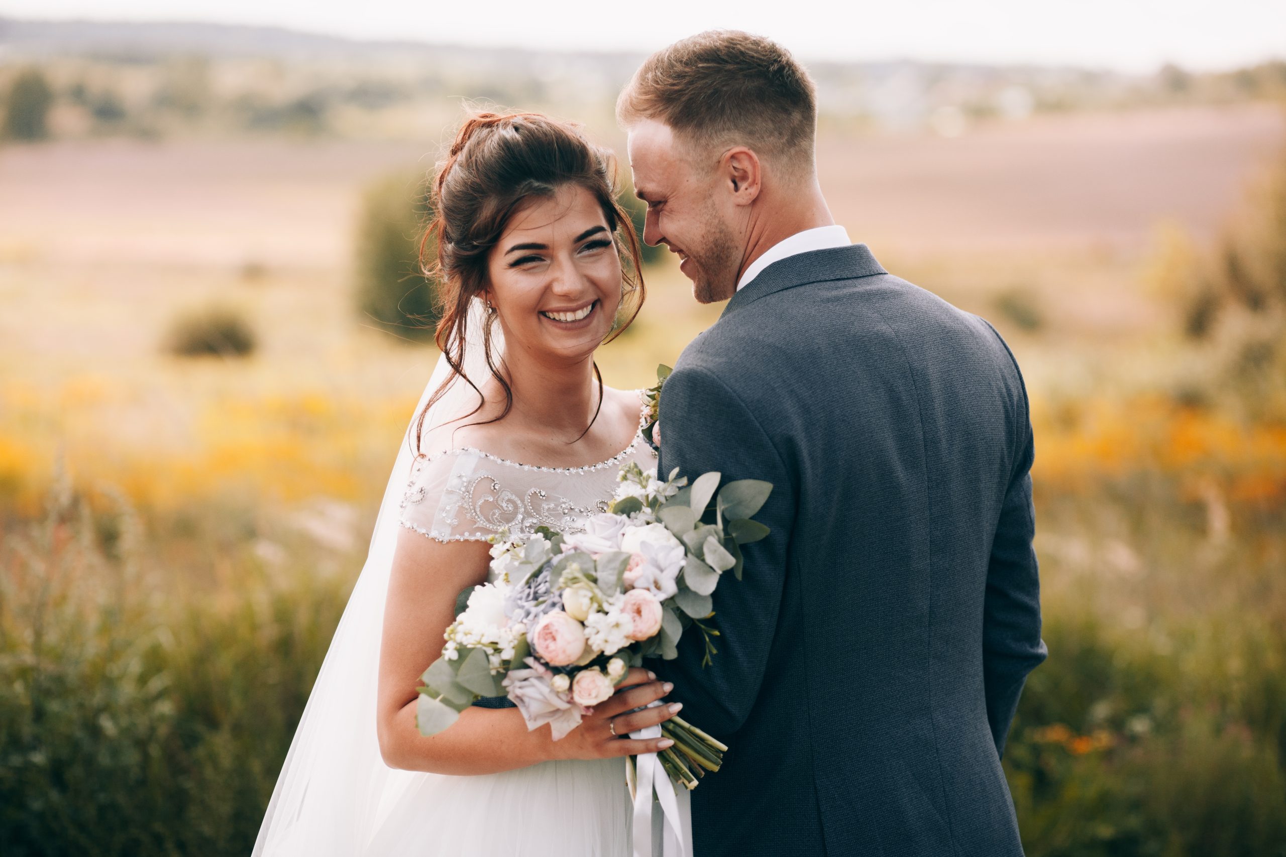 A smiling woman in a white wedding dress holds a bouquet and looks at a man in a gray suit, both standing in a field with colorful flowers in the background.