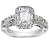 A silver ring with an emerald-cut center stone is surrounded by intricate detailing and smaller stones along the band.