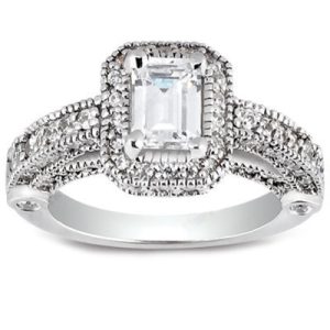 A silver ring with an emerald-cut center stone is surrounded by intricate detailing and smaller stones along the band.
