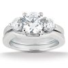 A three-stone engagement ring features a large central diamond flanked by two smaller diamonds, set in a polished white metal band.