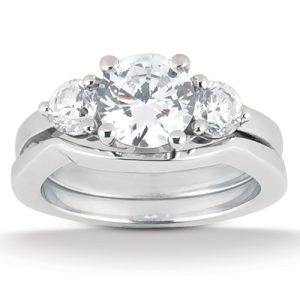 A three-stone engagement ring features a large central diamond flanked by two smaller diamonds, set in a polished white metal band.