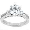 A silver three-stone ring features a large central clear gemstone flanked by two smaller stones, all set on a shimmery band with additional small inlaid stones.