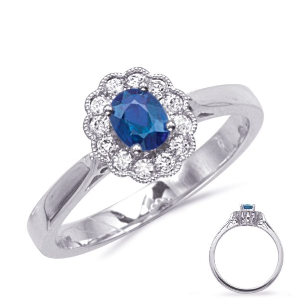 A silver ring with a blue oval gemstone surrounded by a halo of smaller clear stones is displayed at an angle.
