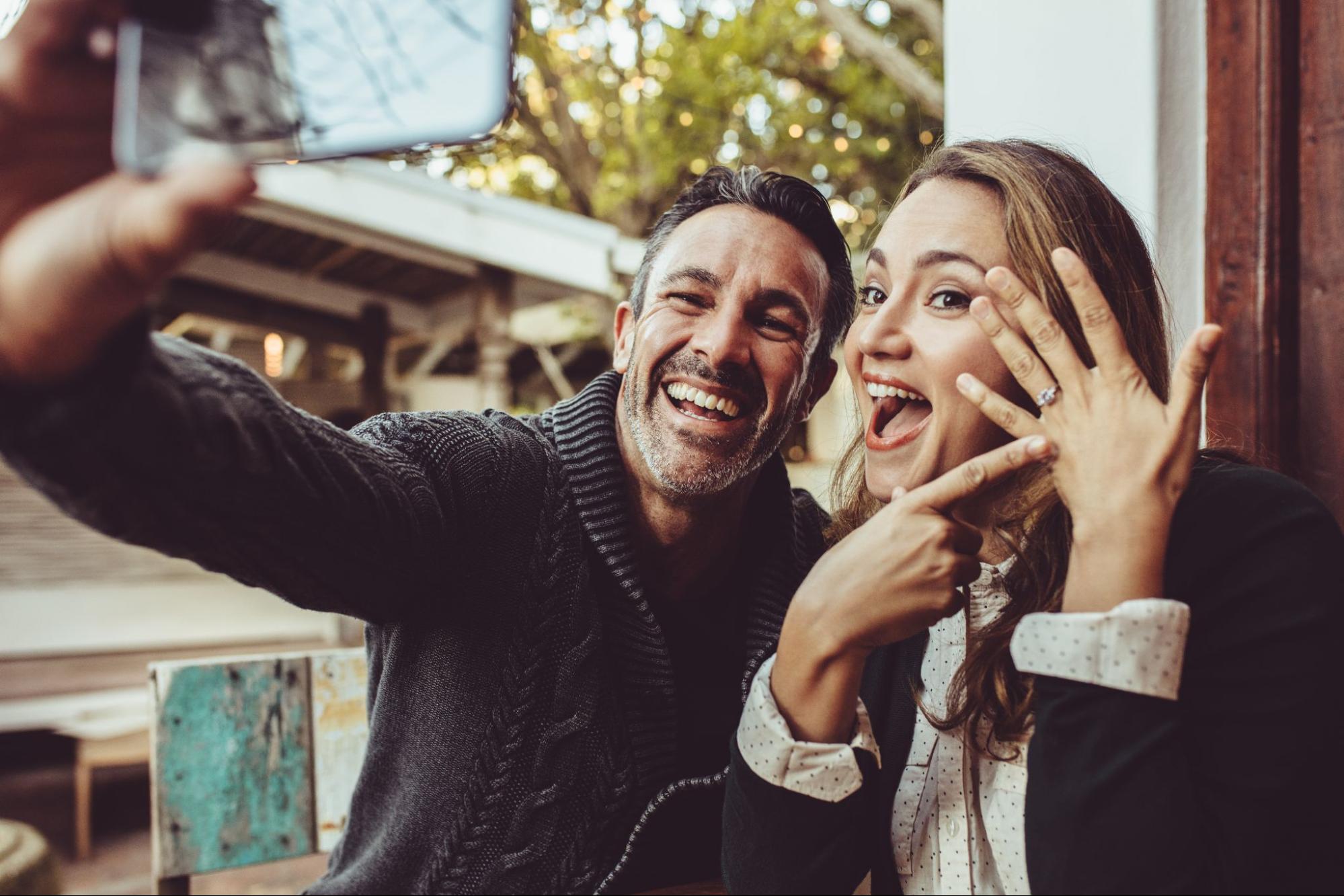 A couple posing for a selfie pointing at an engagement ring